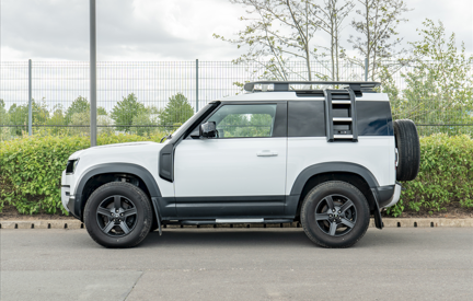 White 4x4 vehicle with a roof rack and spare tyre on the back, parked on a roadside.