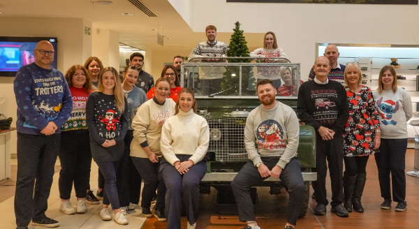 Group of people wearing Christmas jumpers posing around an off-road vehicle indoors.