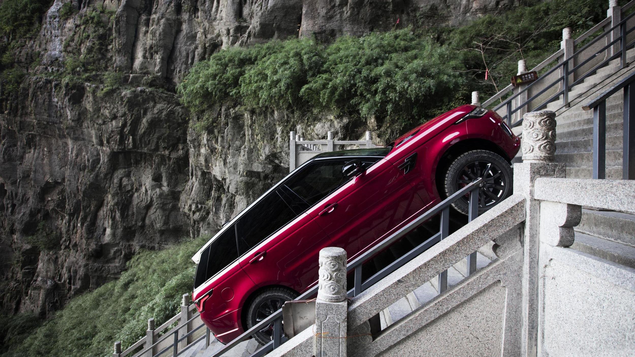 Red SUV ascending steep outdoor stone staircase with rocky cliffside in view.