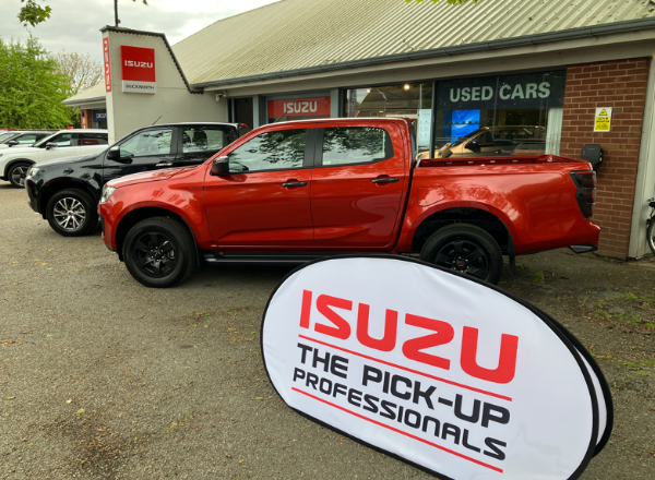 Red Isuzu pick-up truck parked outside a dealership with a promotional sign in front.