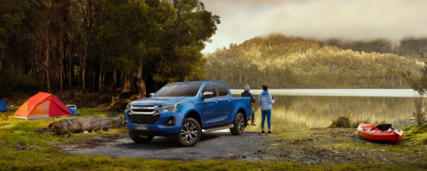Blue pickup truck parked by a lake, with a tent and kayak nearby. Two people look at the water.