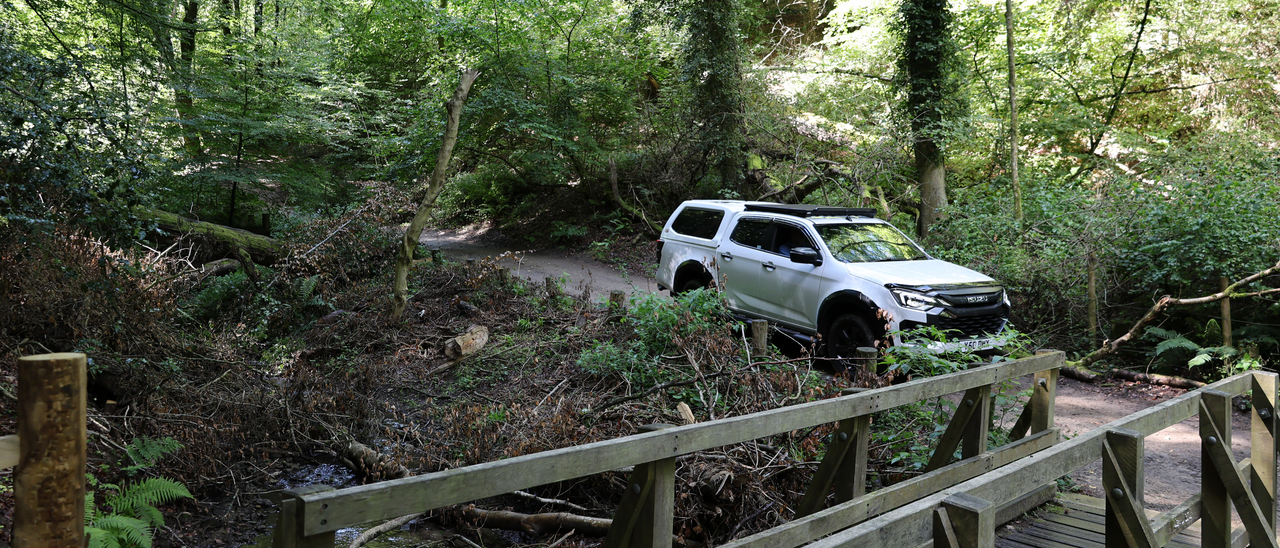SUV parked on a dirt path in a forested area, next to a wooden bridge.