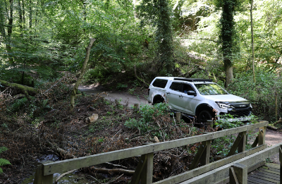 White SUV driving through a forested dirt path beside a wooden bridge.