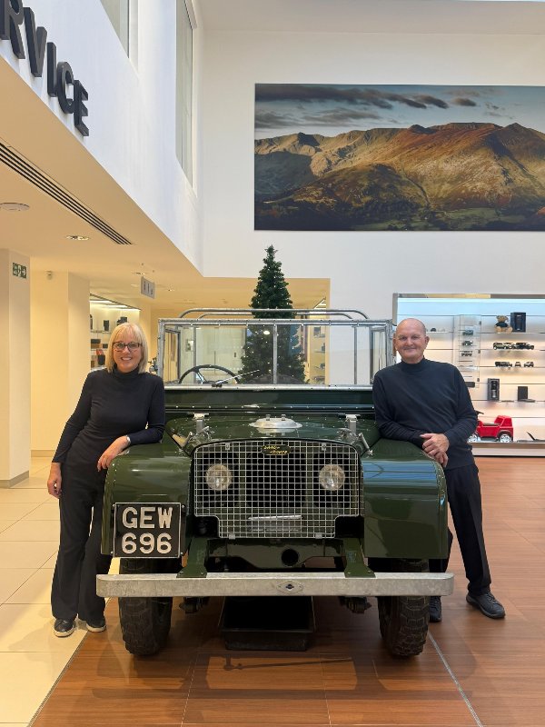 Two people stand beside a vintage green Land Rover in a showroom.