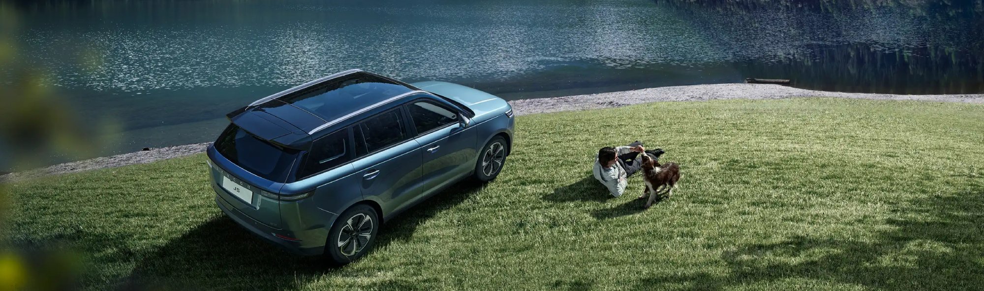 SUV parked by a lake with mountains; man and dog on grass nearby.