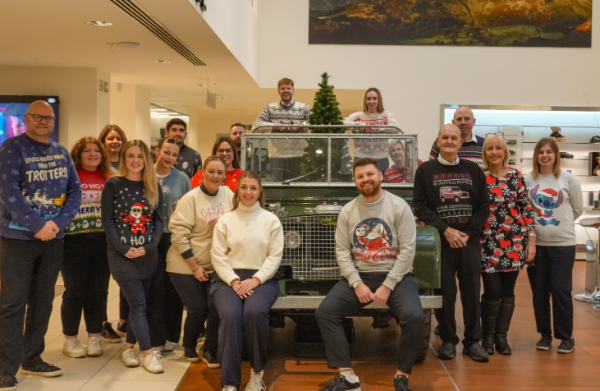 Group of people wearing Christmas jumpers gathered around an old green SUV indoors.