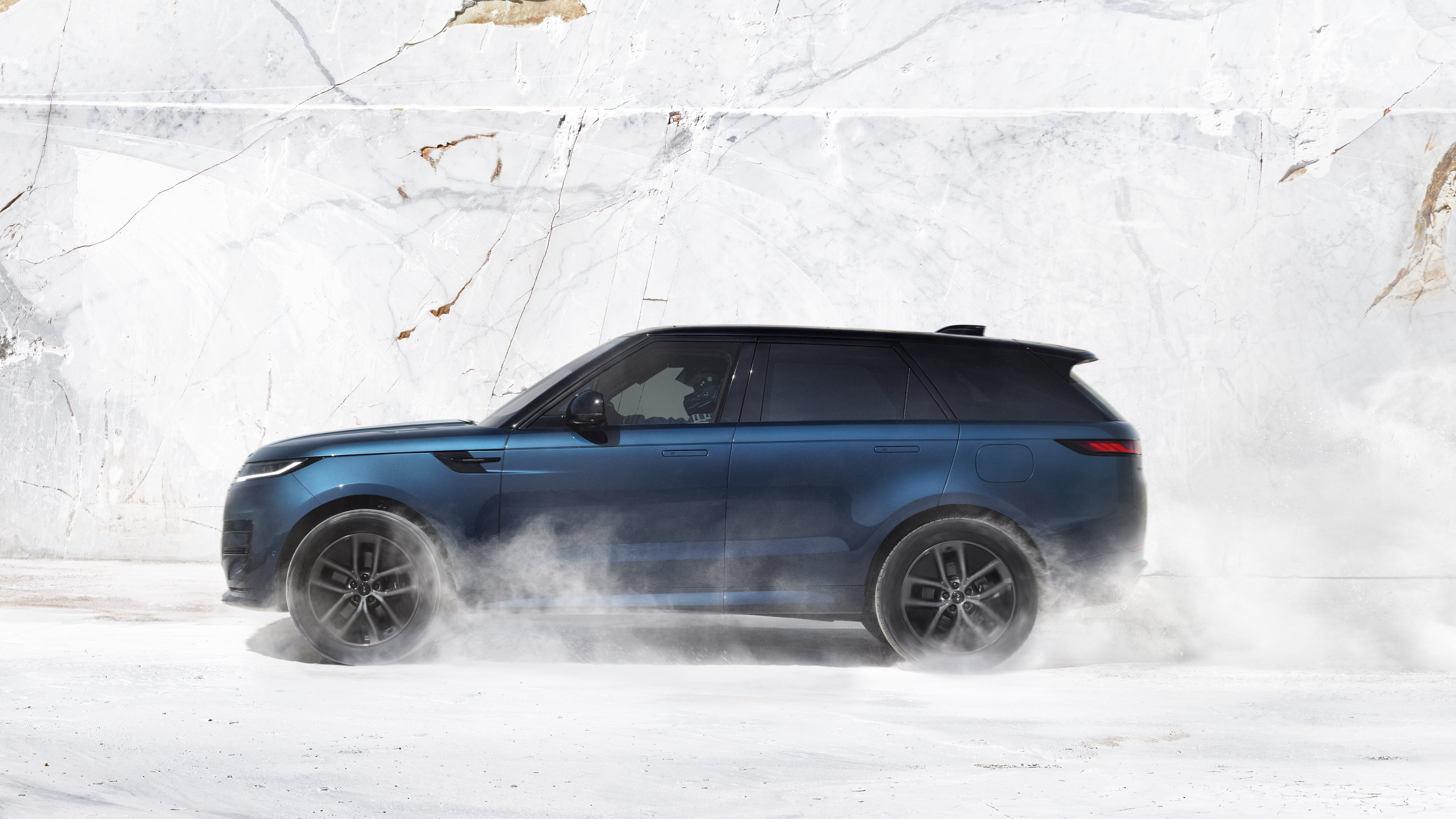 Blue SUV driving on a snowy surface with dust clouds around the wheels.