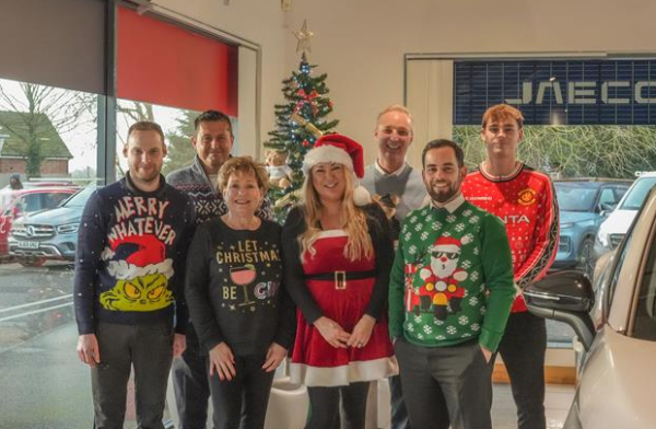 Group of eight people wearing Christmas jumpers, standing next to a decorated Christmas tree.